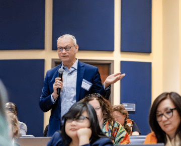 A man in a blue suit jacket and glasses holds a microphone and gestures with his left hand while asking a question at a panel.