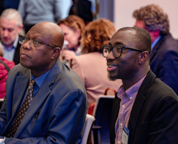 Two men in suits and glasses intently listen at a session with other people in the background.