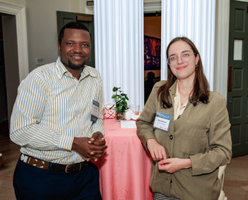 Two individuals stand next to a table with a pink tablecloth. The individual on the left wears a striped shirt and dark pants. The individual on the right wears glasses and a light brown jacket.