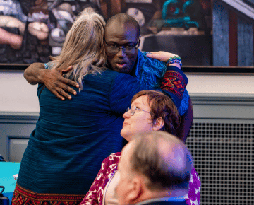 A man in glasses wearing a blue shirt  hugs a person with a blue shirt and colorful trim.