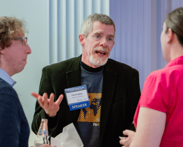 A man with graying hair and a beard gestures with his hands while speaking to two other people. He wears a dark jacket over a t-shirt with a graphic design and a name tag.