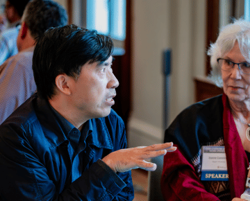 A man in a dark jacket gestures with his hands while speaking to a woman wearing glasses and a conference name tag.