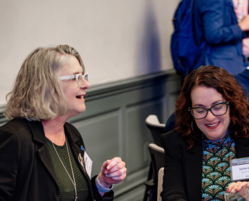 Two individuals are engaged in conversation, one with gray hair and glasses, wearing a black blazer, and the other with curly brown hair and glasses, also wearing a black blazer.