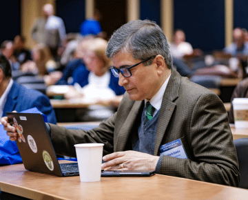 A person wearing glasses and a brown jacket sits at a table during a session with a laptop and a white cup.