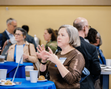 A woman with gray hair gestures with her hands while speaking at a lunch. Several other people are visible in the background.