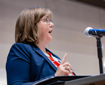 A woman in a navy blazer and red, white, and blue shirt speaks into a microphone at the opening plenary.