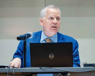 A man in a blue suit and tie speaks into a microphone at a table with a laptop.