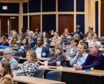 A large group of conference attendees sit in an auditorium, seated at long tables facing forward.