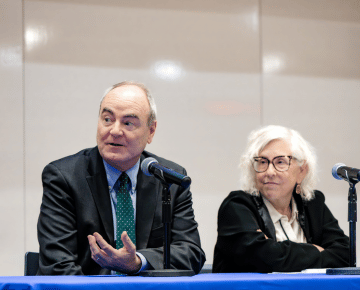 A man in a suit and tie speaks into a microphone, while a woman in glasses sits beside him at a conference panel.