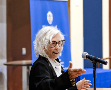 A person with white curly hair and glasses speaks animatedly with their hands outstretched, standing behind a microphone with a blue banner in the background.